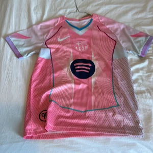 Rosa FC Barcelona Nike fotbollströja - Säljer en limited edition FC Barcelona fotbollströja från Nike i rosa och ljusa toner. Tröjan har korta ärmar, V-ringad krage med blå kant, vita och mörkrosa detaljer samt klubbmärke och Spotify-logga på bröstet. Tillverkad i Dri-Fit material för bästa komfort.