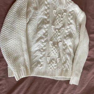 Vit stickad tröja med flätmönster - Säljer en supermysig vit stickad tröja med grovt flätmönster och hög ribbad krage. Tröjan har långärmade ärmar och är perfekt för kyliga dagar. Den har en klassisk look som aldrig går ur tiden.