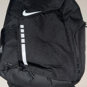 Svart Nike Elite ryggsäck - Helt ny snygg svart Nike Elite ryggsäck med vita detaljer och logga. Väskan har flera fack, justerbara axelremmar med vaddering och transparenta detaljer. Tillverkad i slitstark polyester och perfekt för att bära allt du behöver till skolan eller träningen.