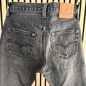 Levi's 501 svarta jeans W27 L30 - Snygga svarta Levi's 501 jeans med klassisk rak passform och fem fickor. Jeansen har en cool tvättad look, knappgylf och detaljer i kopparfärg. Perfekta för dig som gillar tidlös stil och vill ha ett par jeans som funkar till allt.