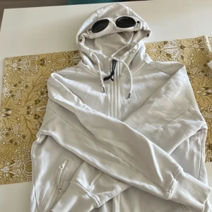 Vit hoodiejacka med goggles från C.P. Company - Vit hoodiejacka från C.P. Company med ikoniska goggles i huvan. Jackan har dragkedja framtill, snörning i huvan och ribbade muddar. Tillverkad i mjuk bomull och har praktiska fickor med dragkedja. Perfekt för dig som vill sticka ut med streetwear-vibe. Jag är 185 lång, har passat perfekt. Inga fläckar. Lite sliten i muddar men inget märkbart. 