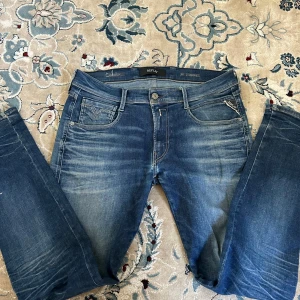 Replay Anbass Hyperflex jeans blå - Säljer ett par Replay Anbass Hyperflex jeans i klassisk blå tvätt med snygga slitningar. Jeansen har fem fickor, normal passform och stretchigt material för extra komfort. Perfekta för dig som gillar stilrena och bekväma jeans med modern look.