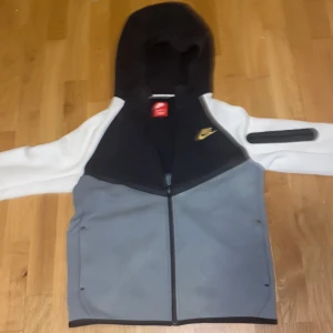 Nike hoodie jacka i svart, grå och vit XL - Nike hoodie jacka i färgblockad design med svart huva, grå front och vita ärmar. Jackan har hel dragkedja, lång ärm och en liten ficka med dragkedja på ena ärmen. Perfekt för en sportig och chill stil.