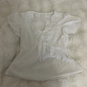 Vit blus med spets och broderi - Söt vit blus med v-ringning och spetsdetaljer längs halsen. Blusen har korta ärmar och är dekorerad med broderade hålmönster över hela tyget. Den har en lätt utsvängd passform och romantisk känsla, perfekt för ljusa dagar.