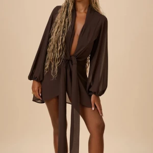 Brun klänning Freia - Snygg brun kort omlottklänning med djup v-ringning och långa, luftiga ärmar. Klänningen har ett brett knytband i midjan som ger en markerad siluett. Perfekt för dig som vill ha en trendig och chic look.