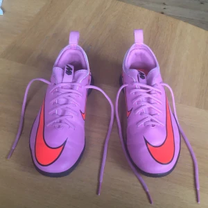 Rosa Nike inne fotbollsskor med orange swoosh - Säljer ett par riktigt snygga rosa Nike fotbollsskor med stora orange swoosh-loggor på sidorna. Skorna har svart sula med bra grepp och klassisk snörning. Perfekta för dig som vill sticka ut på planen och köra din egen stil.