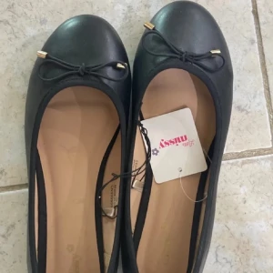 Svarta ballerinaskor med rosett - Snygga svarta ballerinaskor från Missy med rund tå och dekorativ rosett framtill. Skorna har guldfärgade detaljer på rosetterna och en klassisk, stilren design. Perfekta för dig som gillar enkel och tidlös stil.