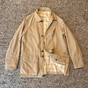 loro piana horsey jacket - Stilren beige jacka från Loro piana med klassisk krage, stora fickor och rutigt innerfoder. Jackan har både dragkedja och knappar framtill för extra funktionalitet. Perfekt för dig som gillar en clean och tidlös look.