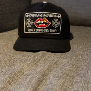 Svart Chrome Hearts keps med mesh - Säljer en svart keps från Chrome Hearts med mesh på sidorna och bak, samt justerbar snapback. Framtill finns en stor broderad patch med text, vita kors och röda läppar. Metallknapp med logga på toppen. Riktigt street och unik stil.