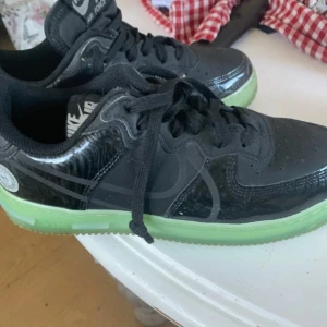 Nike Air Force 1 All Star Svart/Grön - Säljer ett par Nike Air Force 1 All Star med svart ovandel i glansigt syntetmaterial och coola detaljer. Sulan är i en fräsch ljusgrön färg som verkligen sticker ut. Skorna har snörning och rund tå, samt en specialdetalj med All Star-logga på hälen.