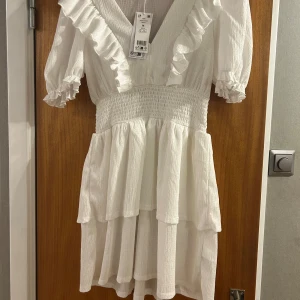Vit smockad volangklänning M - Superfin vit kortklänning med smockad midja, v-ringning och volanger längs ärmar och kjol. Klänningen har korta puffärmar med resår. Helt ny, endast testad. Hade passat perfekt som studentklänning!💗 Nypris: 399 kr