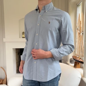 Ljusblå skjorta från Ralph Lauren - Säljer en stilren ljusblå skjorta från Ralph Lauren med klassisk button-down krage och broderad logga på bröstet. Skjortan är i bomull och har lång ärm samt custom fit-passform. Perfekt för dig som gillar en clean och tidlös look.