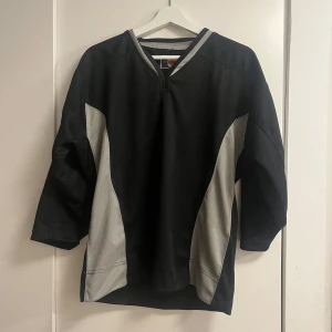 Svart och grå hockeytröja Sher-Wood - Säljer en svart och grå hockeytröja från Sher-Wood med V-ringad hals och trekvartsärm. Tröjan har sportig design med kontrasterande grå paneler på sidorna och runt halsen. Perfekt för dig som gillar hockey eller vill ha en cool oversized look.