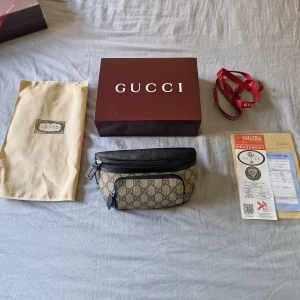 Gucci beige midjeväska med GG-mönster - Snygg beige midjeväska från Gucci med klassiskt GG-mönster och svarta skinn-detaljer. Väskan har flera fack med dragkedja och justerbart band. Perfekt för att bära det viktigaste och ge din outfit en lyxig touch.