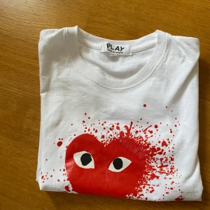 Vit Comme des Garçons Play t-shirt S - Vit t-shirt från Comme des Garçons Play i bomull med korta ärmar. På bröstet finns det ikoniska röda hjärtat med ögon och färgstänk, vilket ger en cool och lekfull vibe. Perfekt för dig som gillar streetstyle och vill sticka ut. Storlek s, (obs trycket har några märken på sig som man kan se på bilderna)
