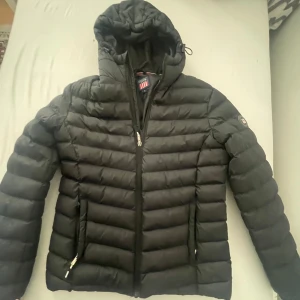 Svart pufferjacka från Vinson, S - Säljer en svart pufferjacka från Vinson i storlek S. Jackan har huva, två fickor med dragkedja och quiltad design. Perfekt för kyliga dagar och har en clean look med diskret logga på ärmen.