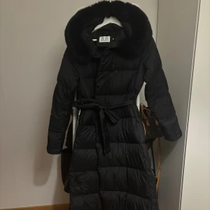 Svart lång dunjacka från FUR Stockholm - Säljer en svart lång dunjacka från FUR Stockholm med fluffig pälskrage och bälte i midjan. 