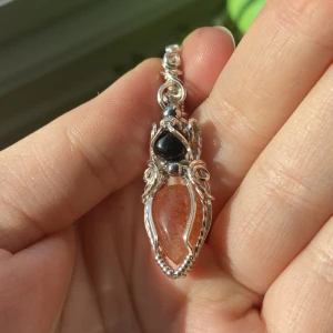 Silverberlock med droppformad sunstone och svart liten sten - Unikt halsband med handvirad silvertråd runt en droppformad orange glittrig sten och en svart sten ovanför. Detaljerad och konstnärlig design med snirkliga former som ger ett magiskt intryck. Perfekt för dig som gillar boho eller fantasy vibes. Kedja i rostfritt stål kostar 50kr.