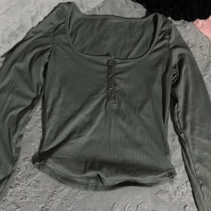 Grå ribbad långärmad topp - Säljer en grå ribbad långärmad topp med rundad, djup urringning och tre knappar framtill. Toppen har en slim passform och är tillverkad i mjukt, stretchigt material som sitter snyggt på kroppen. Storlek är S. Aldrig använt.