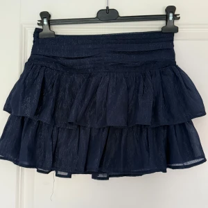 Mörkblå volangkjol från Gina Tricot - Supersöt mörkblå kortkjol från Gina Tricot med flera lager volanger och diskret glittrig effekt i tyget. Kjolen har en markerad midja och är perfekt för dig som vill ha en trendig och lekfull look. Materialet är lätt och ger ett fint fall. Aldrig använd!