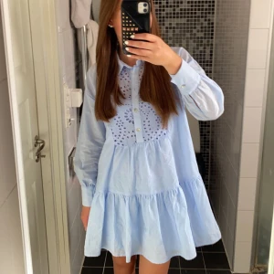Ljusblå volangklänning med broderi - Supersöt ljusblå kortklänning med volanger och broderade detaljer framtill. Klänningen har långärmade ärmar, knappar framtill och en luftig passform. Perfekt för vår och sommar när du vill känna dig extra fin.
