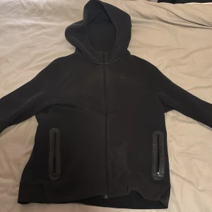 Nike tech hoodie 158-170 cm - Svart hoodie från Nike i storlek XL med hel dragkedja framtill och två fickor med dragkedja. Tröjan har huva och är tillverkad i ett mjukt, stretchigt material som passar perfekt för chill eller träning. Enkel och clean design med diskret Nike-logga på bröstet.