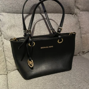 Svart handväska från Michael Kors - Stilren svart handväska från Michael Kors med guldfärgade metalldetaljer och logotyp. Väskan har dubbla handtag, dragkedja upptill och ett rymligt foder med MK-mönster samt flera innerfack. Perfekt för dig som gillar klassisk och lyxig design (ej äkta)