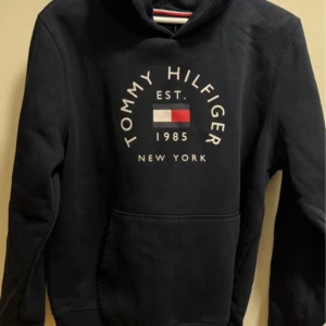 Svart hoodie Tommy Hilfiger - Svart hoodie från Tommy Hilfiger med klassisk logga och text på bröstet. Tröjan har huva, magficka och långa ärmar. Perfekt för en chill och stilren look.
