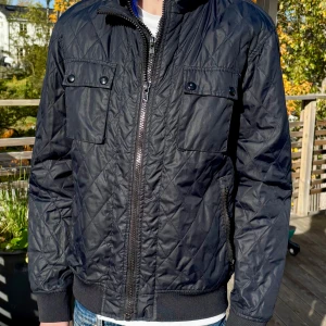 Svart quiltad bomberjacka från Easy Wear - Svart quiltad bomberjacka från Easy Wear med dragkedja framtill, två bröstfickor med knappar och ribbade muddar. Jackan har ett stilrent rutmönster och är tillverkad i syntetmaterial. Perfekt för dig som vill ha en clean och modern look.