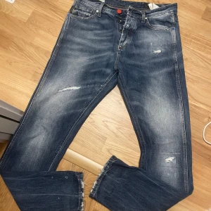 Blå Erik Anti Fit jeans med slitningar - Säljer ett par blå Erik Anti Fit jeans från Nudie Jeans med coola slitningar och råa detaljer. Jeansen har en loose passform och klassisk femficksdesign. Materialet är robust denim i bomull och färgen är snyggt tvättad blå med ljusare partier. 30/32