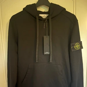 Svart stone island hoodie - Svart hoodie från Stone Island med klassisk logga på vänster ärm. Tröjan har huva med snörning, känguruficka framtill och långa ärmar. Perfekt för dig som gillar streetwear och vill ha en stilren look. Storleken är medium och priset kan diskuteras vid snabb affär😁