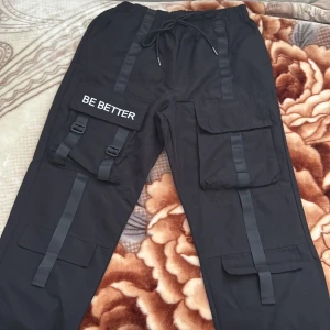 Svarta cargopants med BE BETTER-detalj - Svarta cargopants med elastisk midja och snörning, stora fickor på sidorna och coola banddetaljer. Ena benet har en ficka med texten 'BE BETTER' och spännen. Perfekta för dig som gillar streetstyle och funktionella byxor.