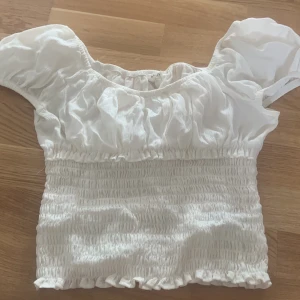 Vit smockad blus med puffärm - Supersöt vit blus med smockad midja och volangig, puffig kort ärm. Toppen har en rynkad struktur upptill och är tillverkad i ett tunt, luftigt tyg. Strl 158 men passar mig som är xxs/xs