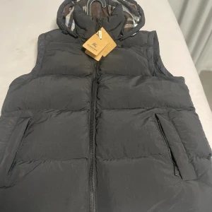 Svart dunväst från Burberry med huva - Svart dunväst från Burberry med dragkedja framtill och två fickor med dragkedja. Västen har en avtagbar huva fodrad med brunt och blått rutigt tyg. Klassisk och stilren design med märkets logga på etiketten. Mycket fin helt ny och är en av burberrys nya design på jackor!