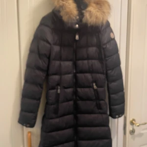 Svart lång dunjacka Moncler med päls - Säljer en svart lång dunjacka från Moncler med fluffig brun päls runt huvan. Jackan har quiltad design, dragkedja framtill och två dragkedjefickor. Perfekt för kalla dagar och riktigt snygg siluett. Klassisk Moncler-logga på ärmen.