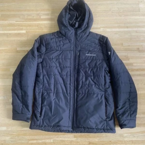 Svart dunjacka Peak Performance - Snygg svart dunjacka från Peak Performance med huva och dragkedja framtill. Jackan har quiltad design, två sidofickor med dragkedja och logga på bröstet samt ärmen. Perfekt för kalla dagar och har en stilren look.