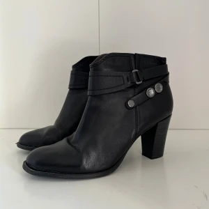 Svarta stövletter från Tamaris - Snygga svarta stövletter från Tamaris i skinn med rund tå och markerad klack. Dekorativa remmar och silvriga knappar på sidan ger en cool detalj. 