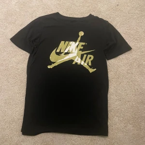 Svart Nike Air Jordan t-shirt guldtryck - Svart t-shirt från Jordan med stort guldigt Nike Air-tryck och Jumpman-logga på bröstet. Klassisk rund hals och korta ärmar. Tillverkad i mjuk bomullsblandning för skön passform. Perfekt för dig som gillar sportig streetstyle.