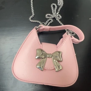 Iconic mini väska🫶 - Söt liten rosa axelväska med kedjerem och en stor silverfärgad rosett framtill. Väskan har en mjuk, rundad form och är perfekt för att lyfta vilken outfit som helst. Materialet är slätt och glansigt, vilket ger en trendig look. Kedjan går att ta av💕