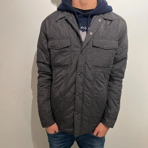 Quiltad grå fieldjacka från J.Lindeberg - Snygg quiltad fieldjacka från J.Lindeberg i mörkgrått. Jackan har tryckknappar framtill, två bröstfickor med lock och raka linjer. Perfekt lager-på-lager-plagg med stilren look och lätt vaddering.