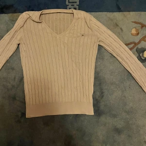 Beige kabelstickad tröja från Gant - Kabelstickad beige tröja från Gant med v-ringning och broderad logga på bröstet. Tröjan har lång ärm och ribbade muddar vid ärmslut och nederkant. Perfekt för dig som gillar klassisk och stilren look.