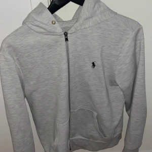 Ljusgrå hoodie från Ralph Lauren - Snygg ljusgrå hoodie från Ralph Lauren med dragkedja framtill och klassisk huva. Tröjan har en broderad logga på bröstet och praktiska fickor. Perfekt för en chill och stilren look.