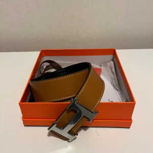 Brunt skärp med H-spänne - Säljer ett snyggt brunt bälte i skinn med ikoniskt H-spänne i silverfärg. Bältet har vita kontrastsömmar och kommer i original orange box med dustbag. Perfekt accessoar för att lyfta din outfit.