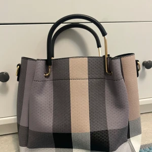 Rutig handväska med svarta detaljer - Snygg handväska med rutigt mönster i beige, svart och grå toner. Väskan har svarta handtag och guldfärgade metalldetaljer. Insidan har flera fack och dragkedja.