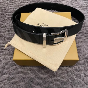 Svart randigt Burberry bälte i skinn - Snyggt svart bälte från Burberry med diskret randigt mönster och silverfärgat spänne. Bältet är gjort i äkta skinn och har en stilren, klassisk design som passar till jeans eller kostym. Perfekt accessoar för dig som gillar exklusiva detaljer och vill sticka ut. Skriv för frågor! Pris kan diskuteras vid snabbköp