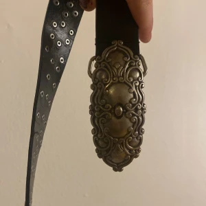 Svart bälte med stor dekorerad silverfärgad spänne - Säljer ett svart bälte i skinn med ett detaljerat silverfärgat spänne. Bältet har inbyggda nitar längs hela remmen som ger en cool och edgy känsla. Perfekt accessoar för att lyfta vilken outfit som helst.
