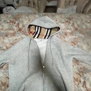 Grå hoodie från Burberry med dragkedja - Säljer en grå hoodie från Burberry med klassiskt rutigt foder i huvan. Tröjan har dragkedja framtill, snörning i huvan och broderad Burberry-logga på bröstet. Tillverkad i mjuk bomullsmix, perfekt för chill dagar.