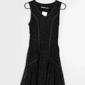 Svart spetsklänning från Desigual - Supersnygg svart ärmlös klänning från Desigual med v-ringning och coolt spetsmönster över hela klänningen. Figurnära upptill och utsvängd nertill, perfekt för dig som gillar unika detaljer och vill sticka ut. Klänningen har ett diskret geometriskt mönster.