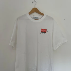 Vit Casablanca Racing t-shirt XL - Casablanca Racing t-shirt i vitt med tryck i rött och svart både fram och bak. Klassisk rund hals, korta ärmar och mjuk bomullskvalitet. Snygg och clean design med tydlig logga och sportig vibe.