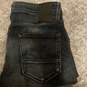 Mörkblå Jack and Jones jeans - Säljer ett par mörkblå jeans med snygga slitningar och blekta partier på benen. Jeansen är i storlek 32 men passar mig som är 180, lite åt de längre hållet för mig till och med.